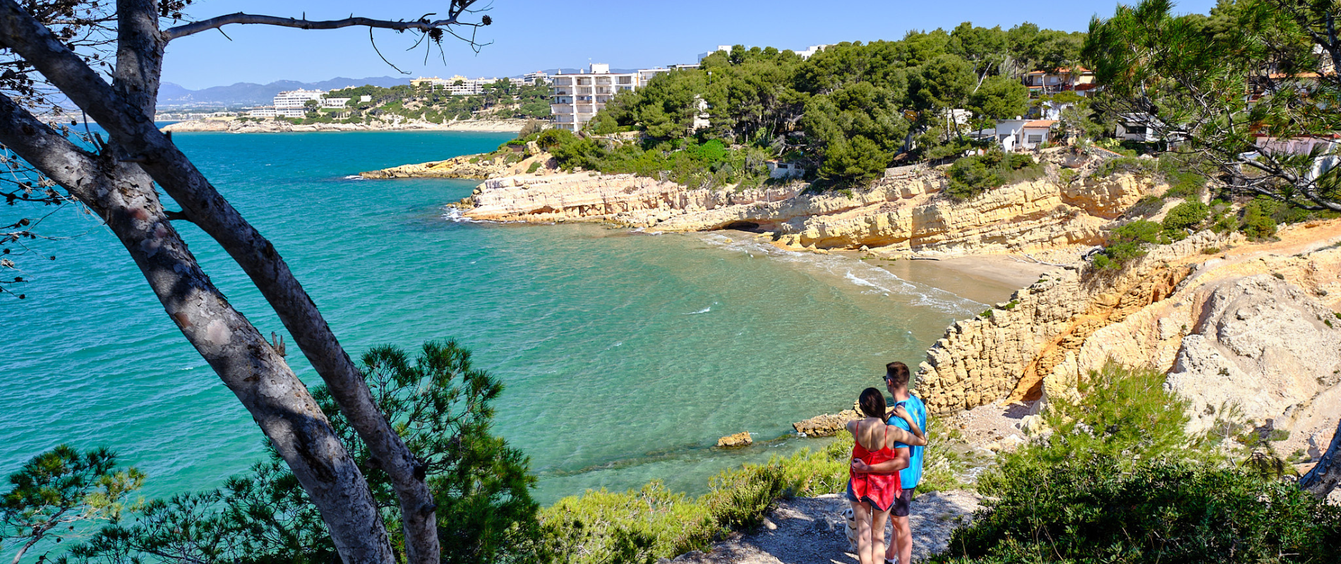 Junges Pärchen steht Arm in Arm auf einem Wanderweg an der Küste und blickt auf der Meer hinaus. Der Küstenstreifen besteht aus Felsen und Strand. Im Hintergrund sind Hauser zu sehen.