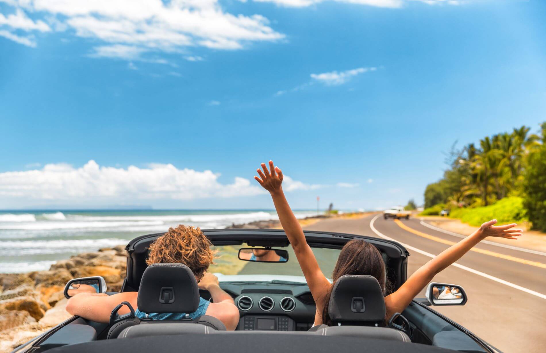 Junges Pärchen im Cabrio im Urlaub auf einer Küstenstraße. Links von der Straße ist das Meer.