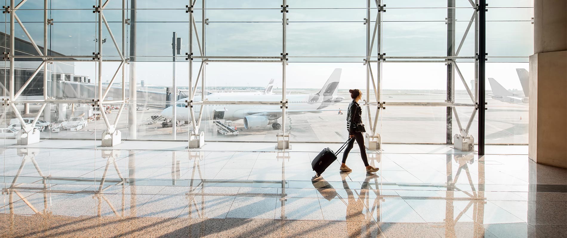 Eine Frau zieht ihren Rollkoffer durch den Flughafen. Im Hintergrund sind große Fenster, durch die mehrere Flugzeuge auf dem Rollfeld zu sehen sind.