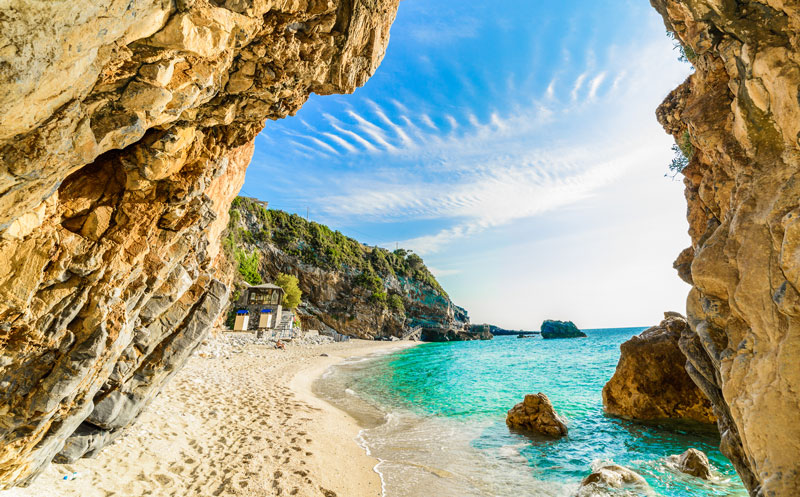 Blick vom Strand auf das azurblaue Meer an einem Sommertag. Links und rechts rahmen Felsen den Blick ein.