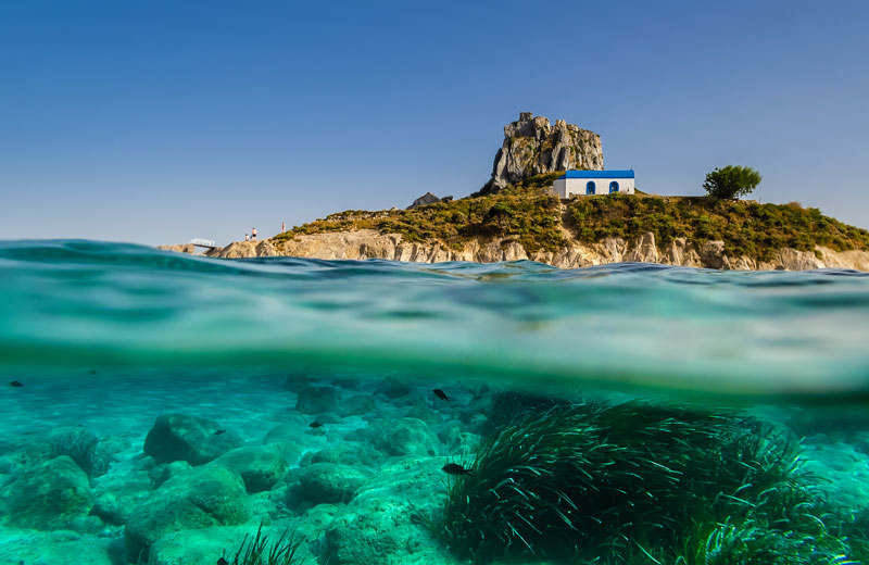 Blick vom Wasser auf den Strand Kefalos an einem schönen Sommertag mit blauen Himmel, an dem ein kleines weißes Haus steht.