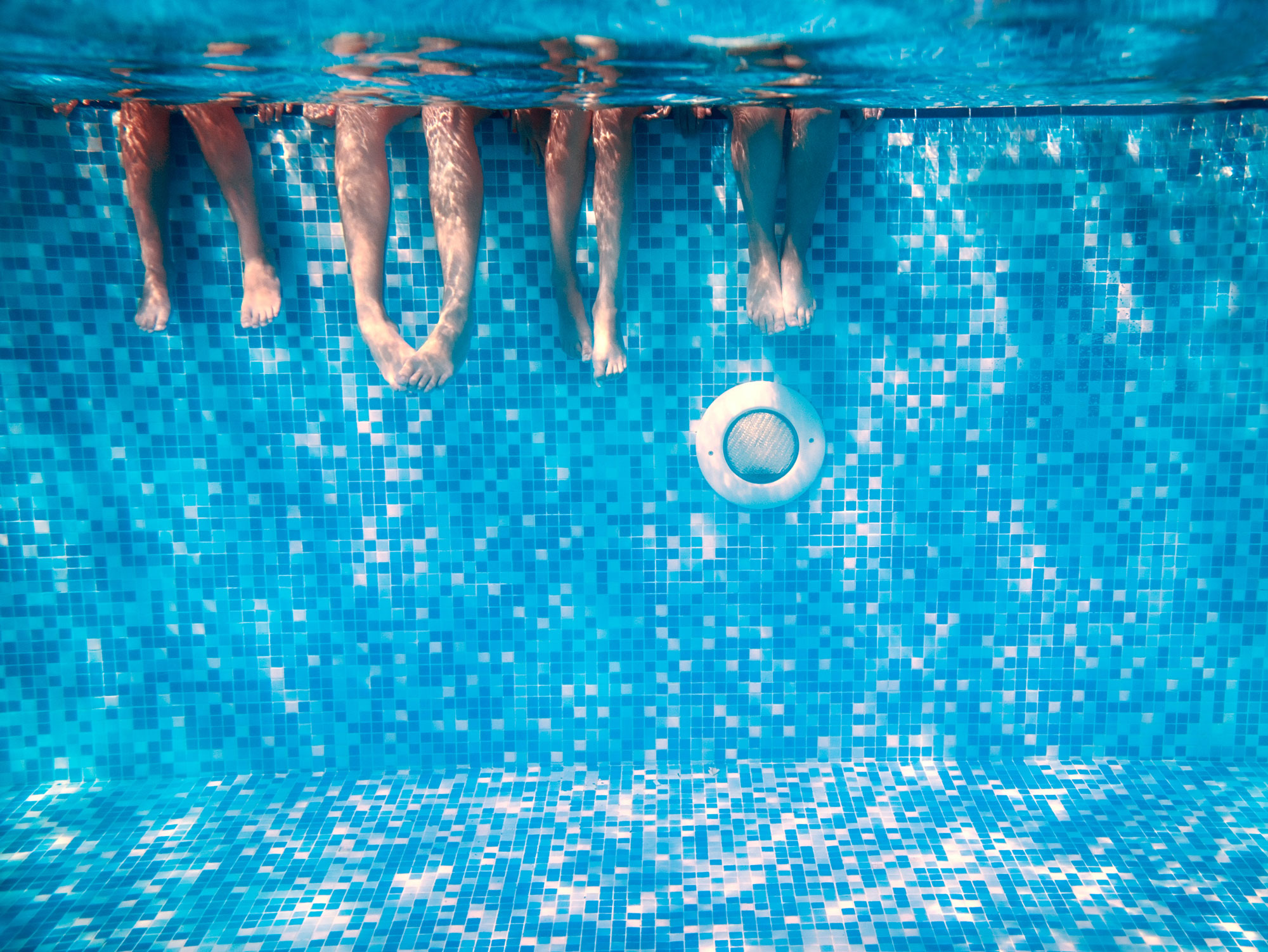 Unterwasseraufnahme in einem Swimmingpool von 4 Paar Füßen von Menschen, die an der Kante sitzen und ihre Beine im Wasser haben.