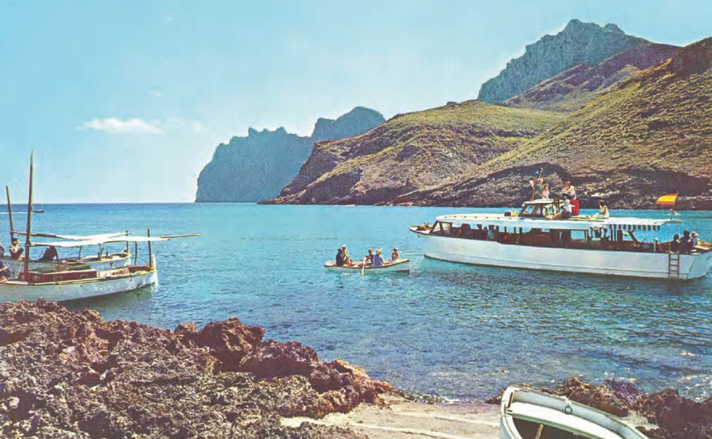 Historische Aufnahme einer Strandszene aus den 60er Jahren auf Mallorca. Zu sehen sind 2 Boote und ein Ruderboot. Das Ruderboot steuert auf eines der beiden Boote zu. Der Strand ist felsig und im Hintergrund sind Berge zu sehen.