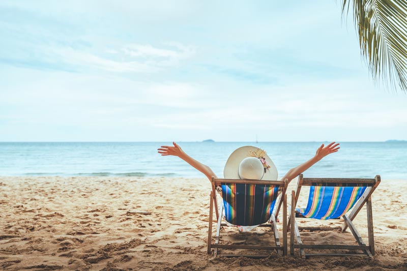 Strand mit zwei Liegestühlen mit Blick auf das Meer. Auf einem sitzt eine Person mit Sonnenhut und streckt die Arme Richtung Himmel aus.