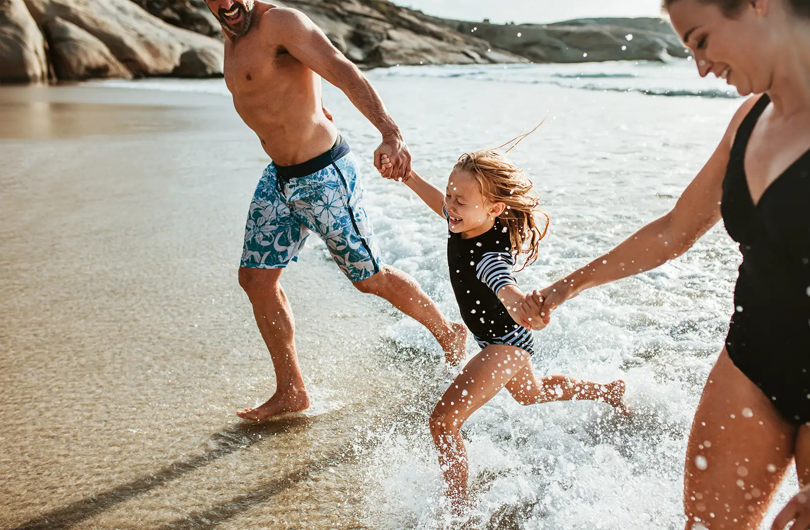 Eltern halten ihre Tochter an den Händen und laufen am Strand aus dem Meerwasser heraus. 