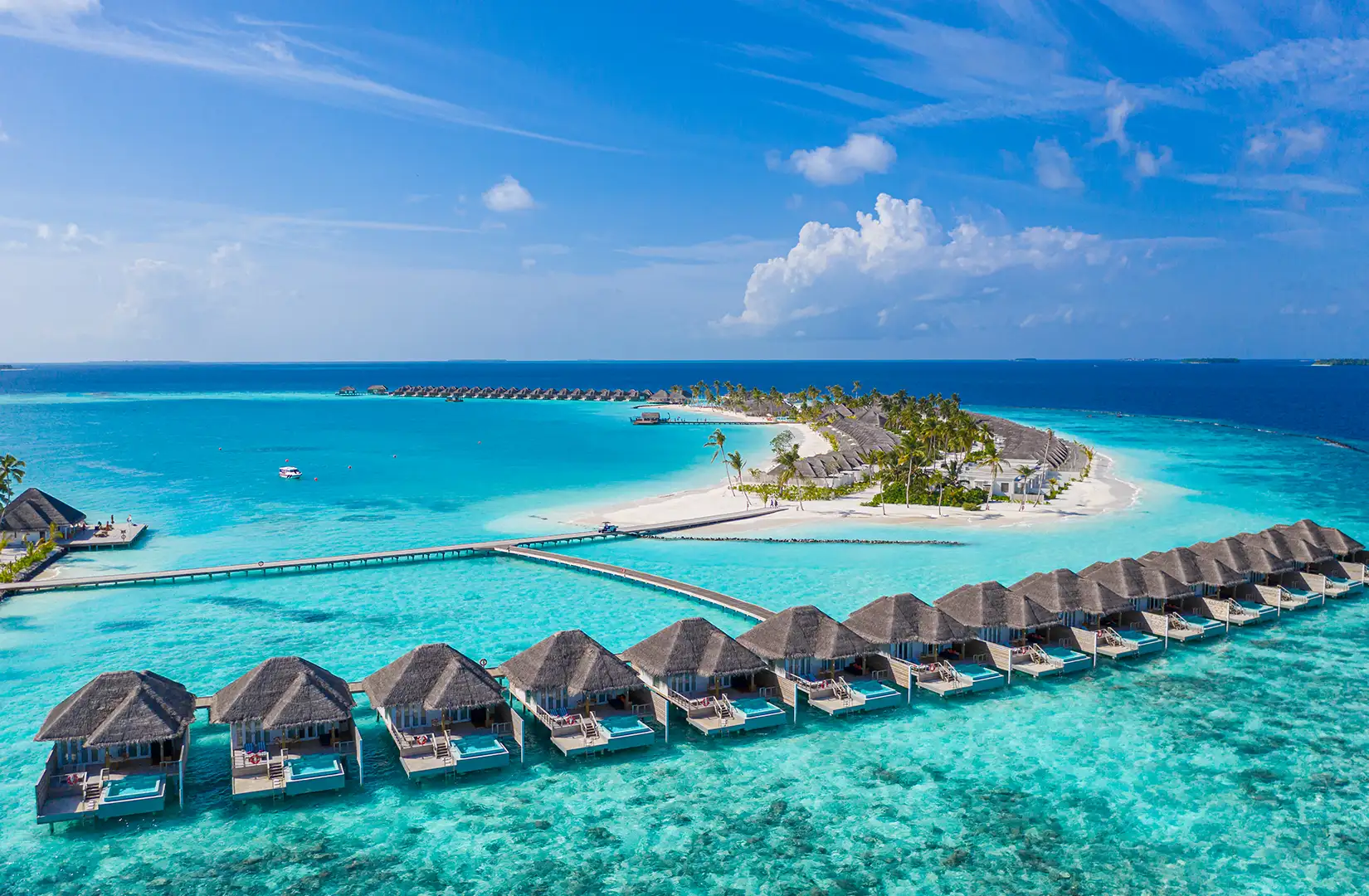 Blick auf eine Malediven-Insel, die mit Hotels bebaut ist bei schönem Wetter. Das Meer, dass die Insel umgibt, ist charakteristisch flach und ist türkisfarben.