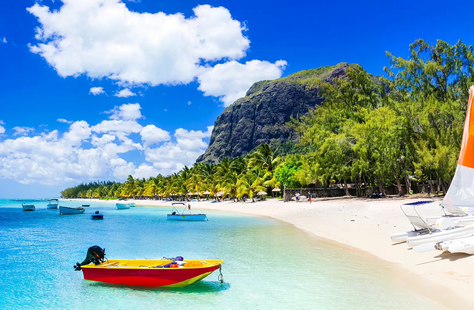 Strand auf der Insel Mauritius an einem Sommertag. Der weiße Strand, rechts, ist von Palmengewächsen gesäumt. Im Wasser anken mehrere kleine Boote.