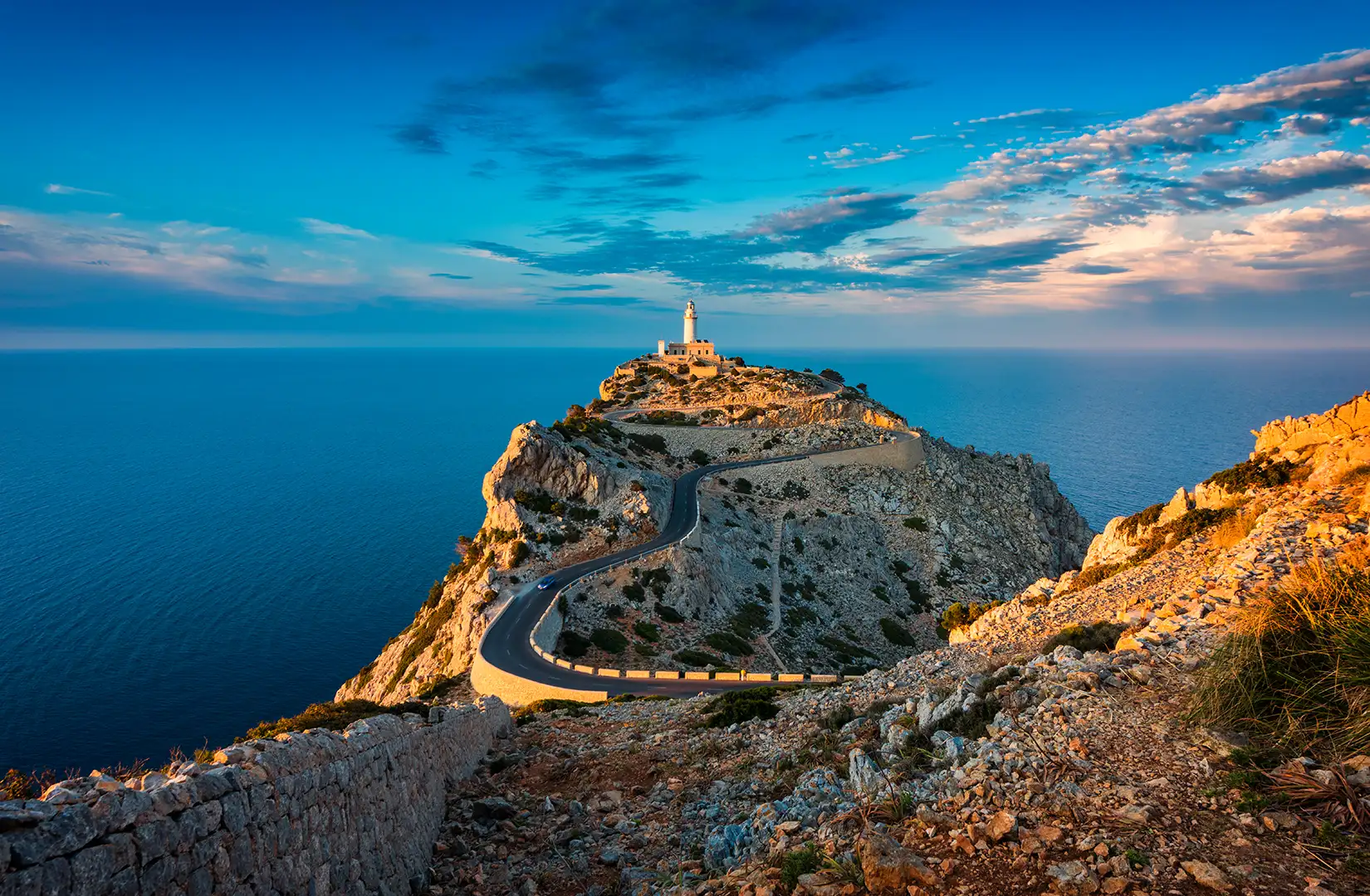 Der berühmte Leuchtturm am Cap de Formentor auf Mallorca bei Abenddämmerung, der einsam auf einem Hügel steht und per Straße angebunden ist.