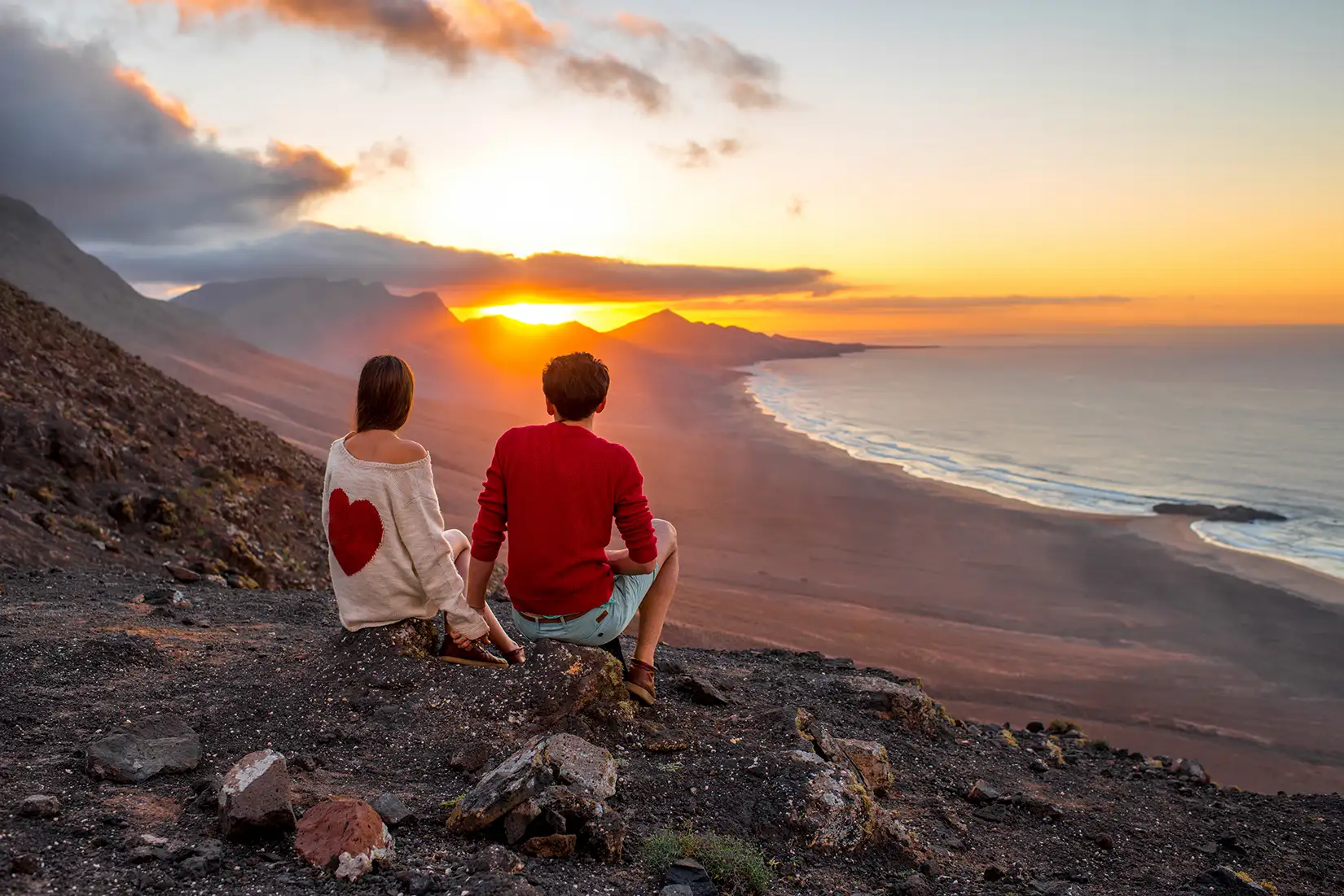 Junges Pärchen genießt die Aussicht auf die Küste von Cofete in einer Sonnenuntergangstimmung auf einem Hügel sitzend mit Blick hinab auf das Meer.