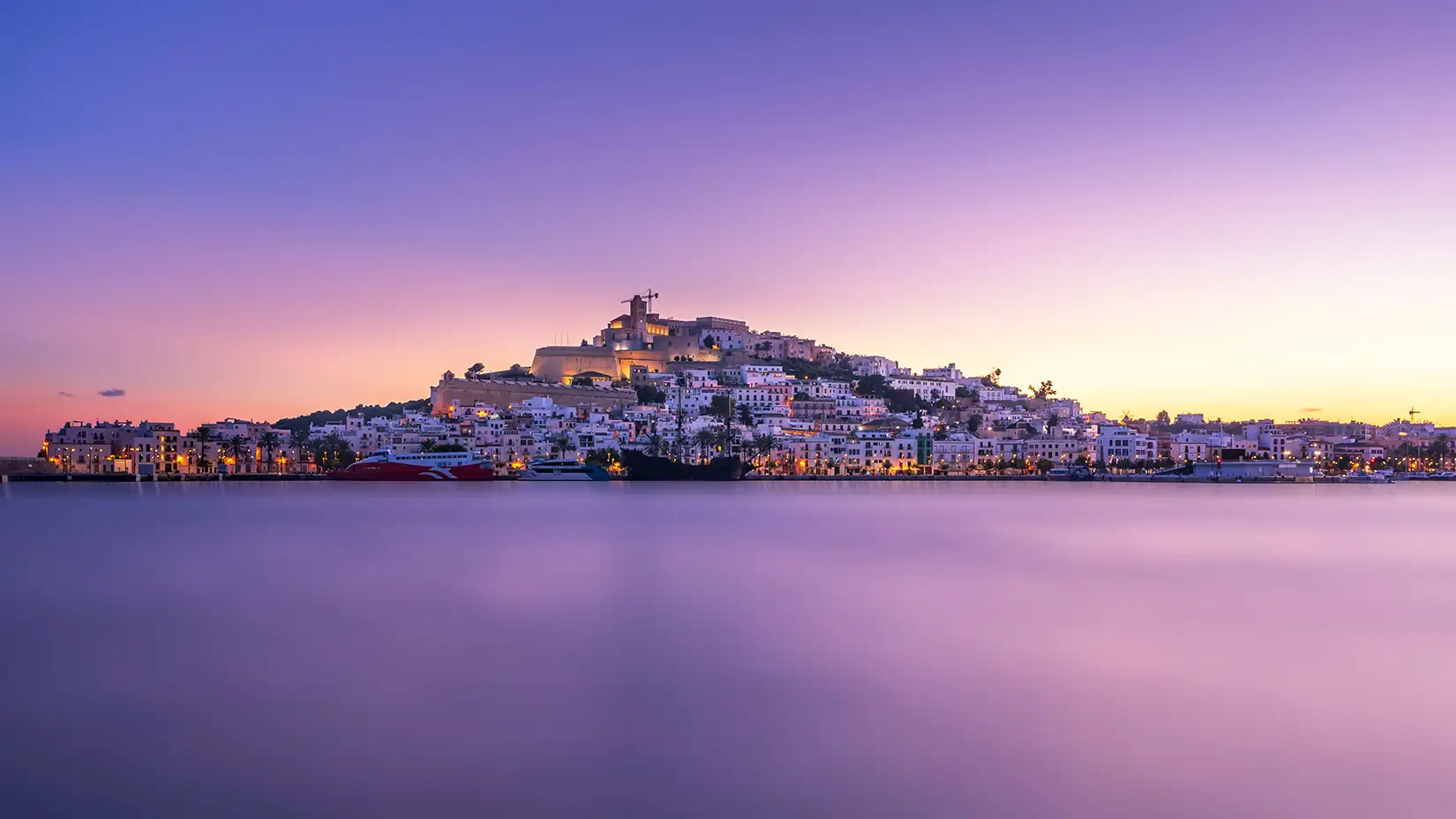Blick auf die Hafenstadt Ibiza vom Wasser ausgesehen in der Abenddaemmerung. 