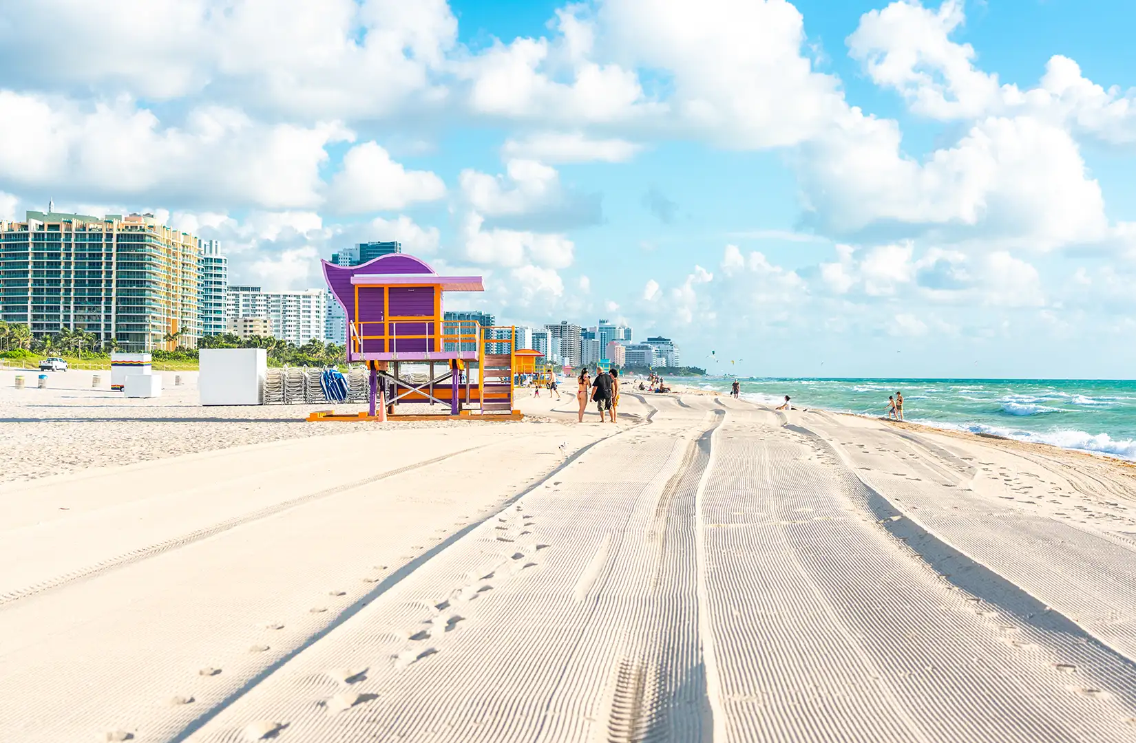 Strandszene mit Rettungsschwimmerhütte und Badenden am Miami South Beach an einem Sommertag. Im Hintergrund schäumen Hotels die Strandlinie.