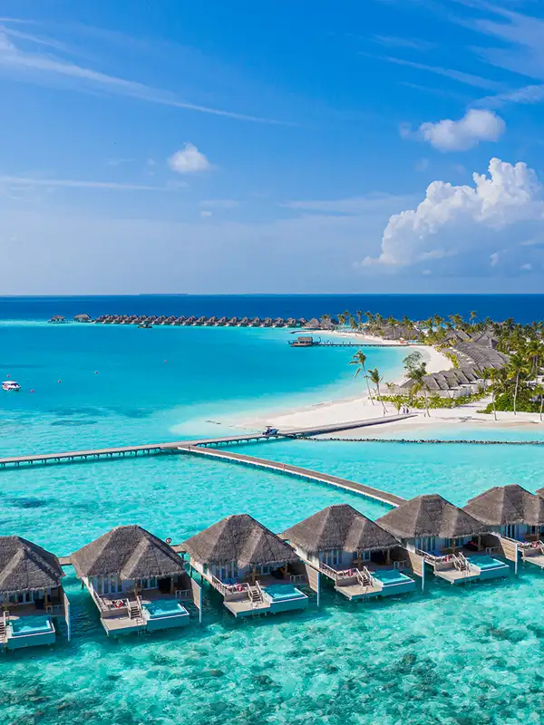 Blick auf eine Malediven-Insel, die mit Hotels bebaut ist bei schönem Wetter. Das Meer, dass die Insel umgibt, ist charakteristisch flach und ist türkisfarben.