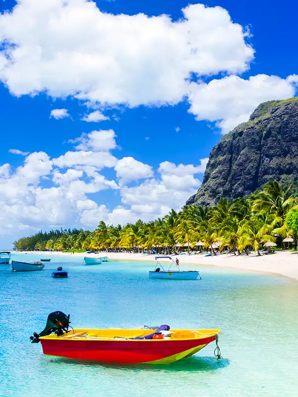 Strand auf der Insel Mauritius an einem Sommertag. Der weiße Strand, rechts, ist von Palmengewächsen gesäumt. Im Wasser anken mehrere kleine Boote.