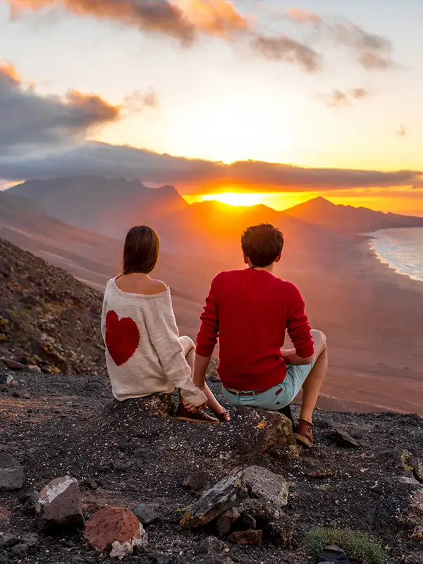 Junges Pärchen genießt die Aussicht auf die Küste von Cofete in einer Sonnenuntergangstimmung auf einem Hügel sitzend mit Blick hinab auf das Meer.