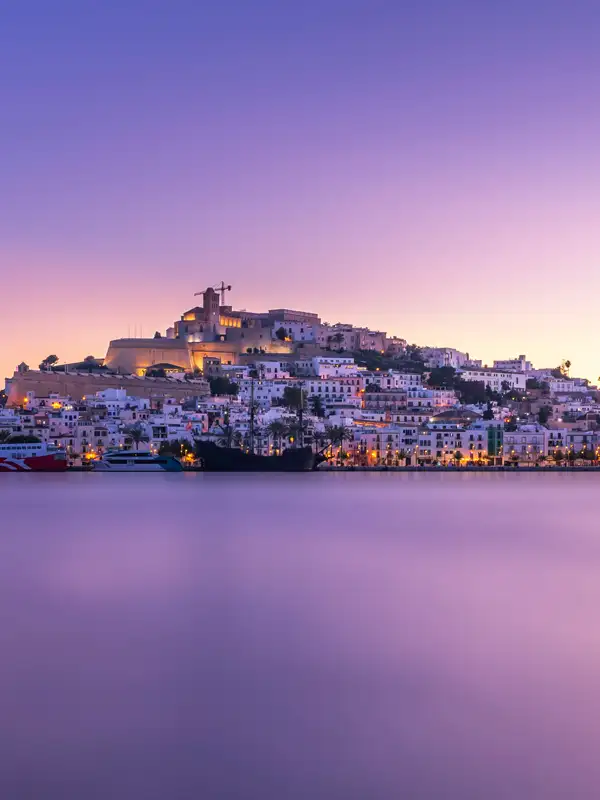 Blick auf die Hafenstadt Ibiza vom Wasser ausgesehen in der Abenddaemmerung. 