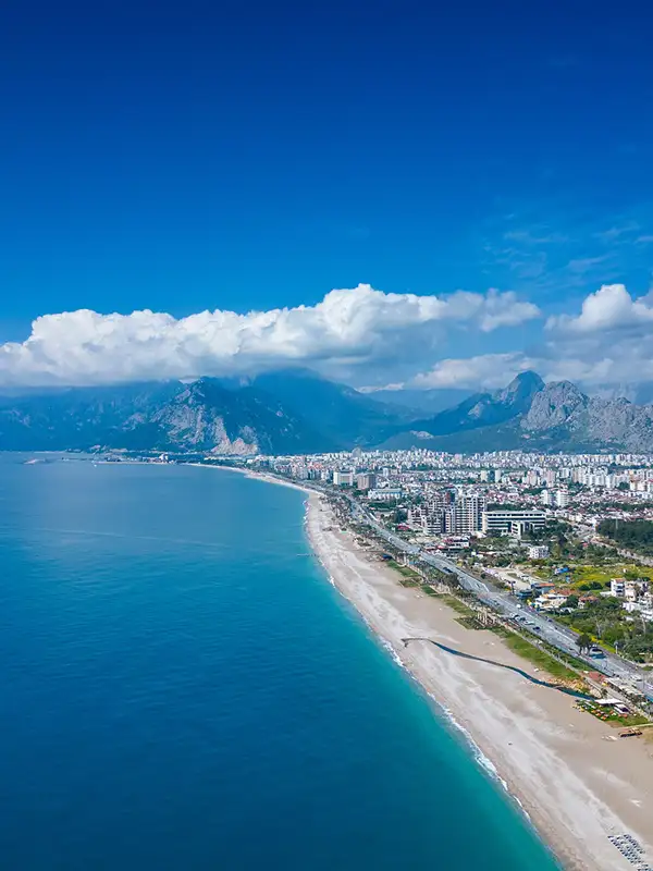 Luftaufnahme von Lara Beach. Das Meer links, rechts der Strand und die dahinterliegende Stadt Antalya. Im Hintergrund ist das wolkenverhangene Gebirge zu sehen.