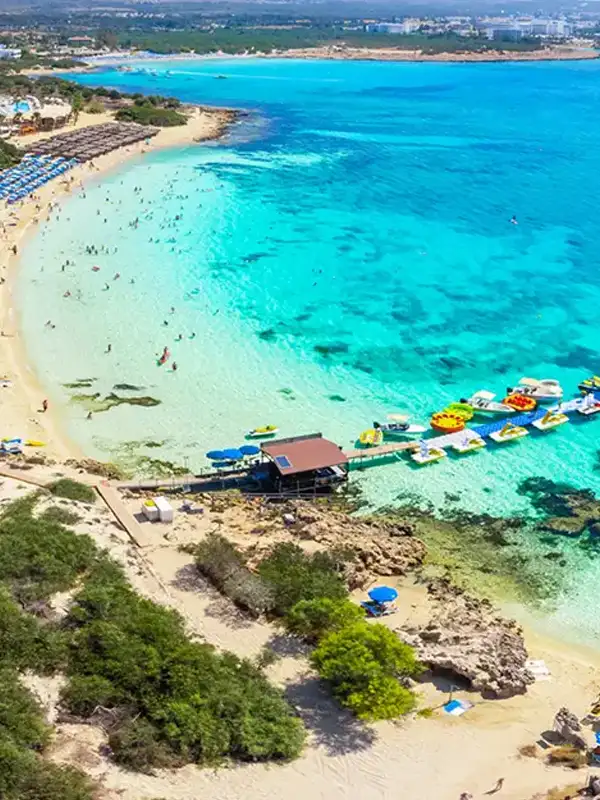 Drohnenaufnahme auf den Sandstrand in der Feigenbaumbucht an einem Sommertag. Die Küste ist mit Hotels bebaut. Am Strand stehen viele Sonnenschirme und Menschen baden im flachen, türkisfarbenem Wasser.