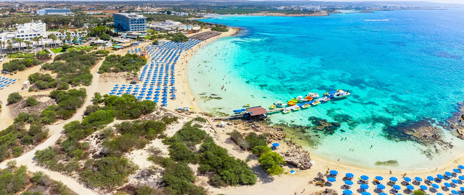 Drohnenaufnahme auf den Sandstrand in der Feigenbaumbucht an einem Sommertag. Die Küste ist mit Hotels bebaut. Am Strand stehen viele Sonnenschirme und Menschen baden im flachen, türkisfarbenem Wasser.